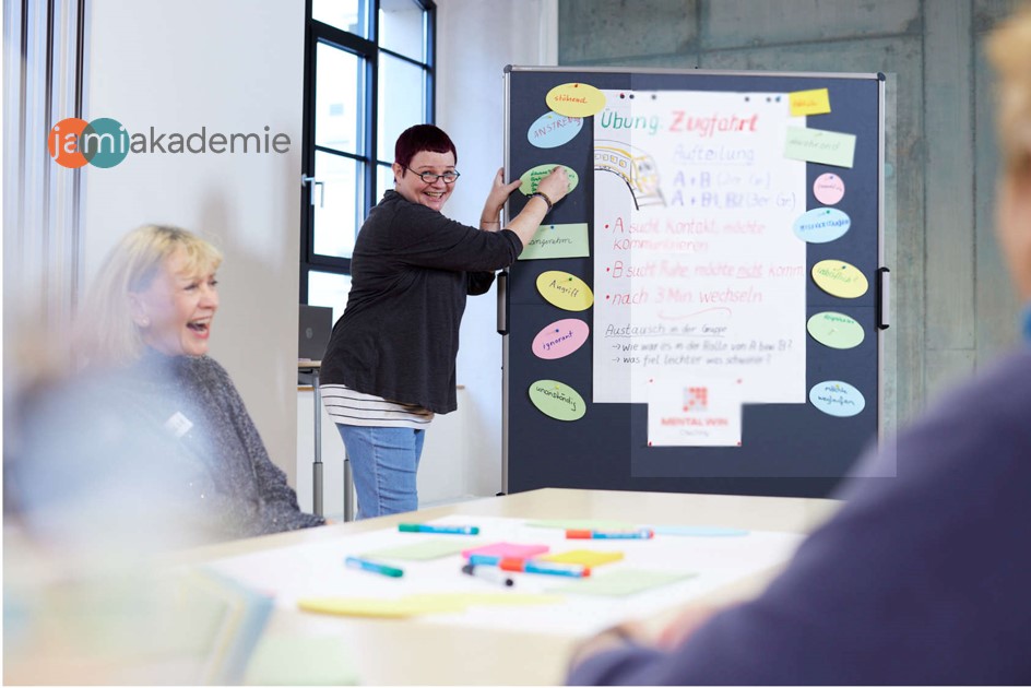 Pinnwand, Person heftet einen Karton an. Lachende Frau im Vordergrund