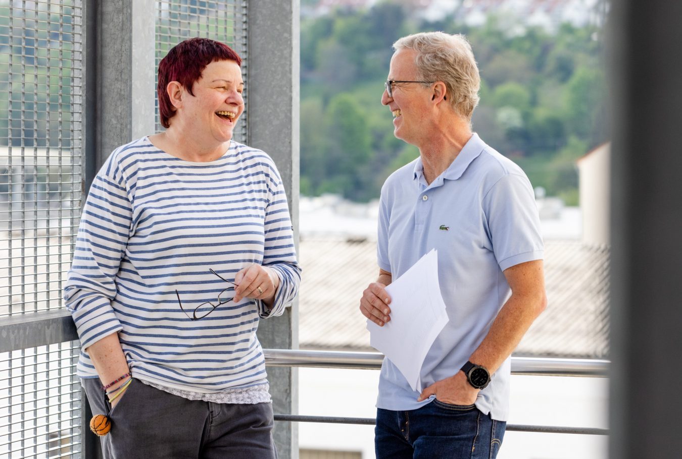 Mann und Frau stehen sich gegenüber und lachen sich an. Frau rechte Hand in der Hostentasche, Mann in rechter Hand einen DinA4 Block