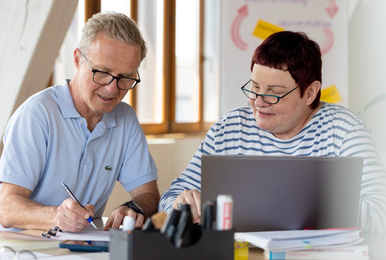 Mann und Frau. Mann schaut auf Papier und zeigt Frau. Frau sitzt vor Laptop