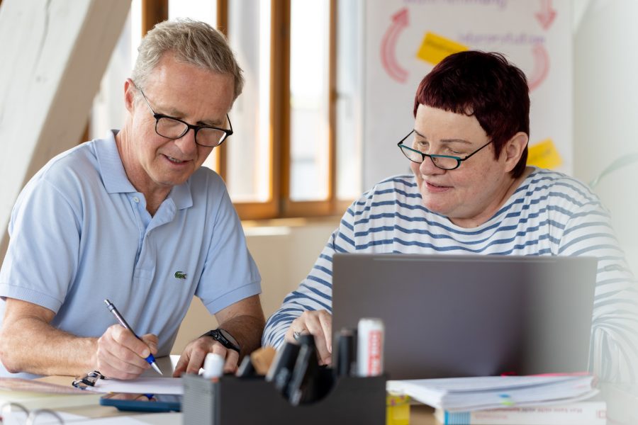 Mann und Frau. Mann schaut auf Papier und zeigt Frau. Frau sitzt vor Laptop