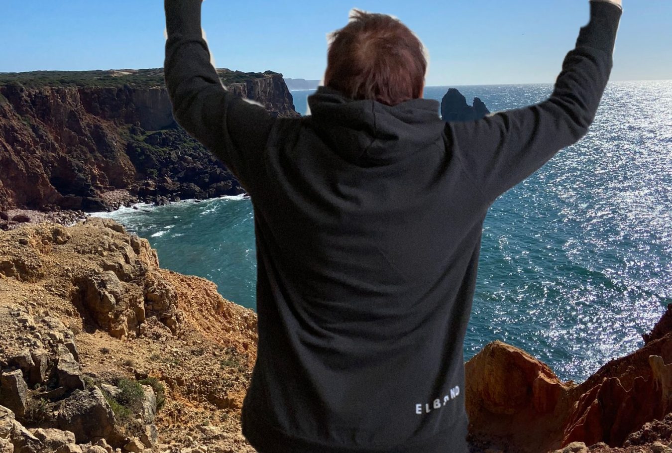 Frau Rücken sichtbar, breitet ihr Arme nach oben aus. Im Hintergrund ist das Meer zu sehen